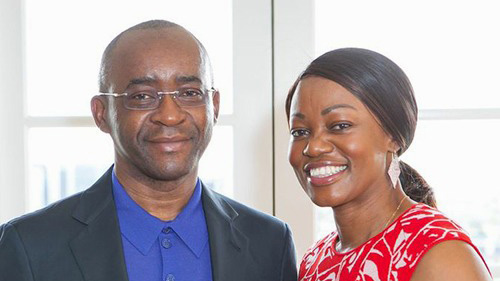 A close up of Strive and Tsitsi Masiyiwa smiling. Strive (left) wears a royal blue polo shirt under a grey suit jacket. Tsitsi (right) wears a red and white patterned shirt with large white earrings.